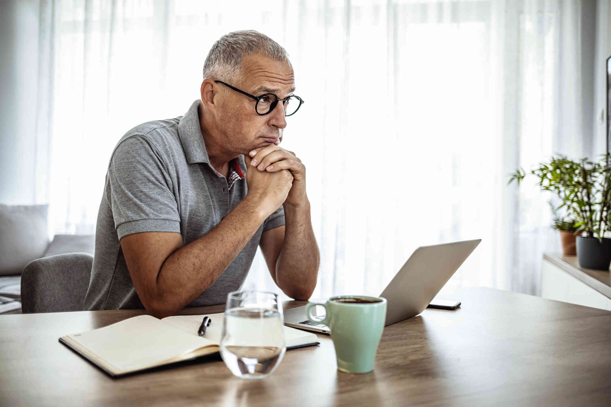 An older man in a grey shirt sits at a desk with a neutral expression while sitting in front of his computer and a coffee mug